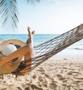Frau mit Strohhut entspannt in einer Hängematte am Sandstrand mit Blick aufs Meer