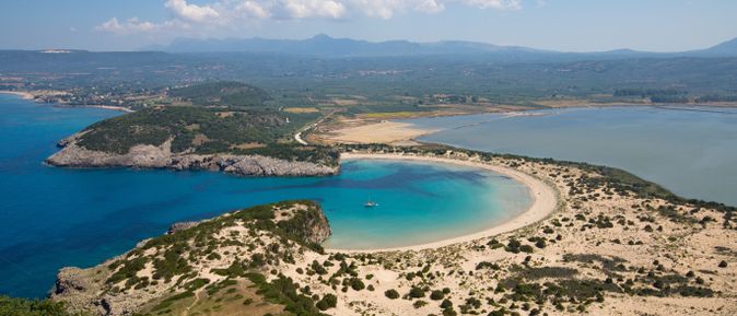 Luftaufnahme einer geschwungenen Sandbucht mit klarem blauem Wasser und bewachsenen Dünen im Vordergrund.