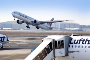 Flughafen Frankfurt mit startender Boeing 777 von Qatar Airways auf der Startbahn, während im Vordergrund ein Lufthansa Airbus A320-200 an einer Fluggastbrücke im Terminal steht