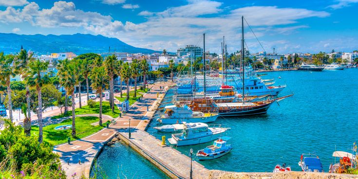 Hafenansicht in Kos, Griechenland, mit mehreren Booten im Wasser, Palmen entlang der Uferpromenade und einem klaren blauen Himmel.