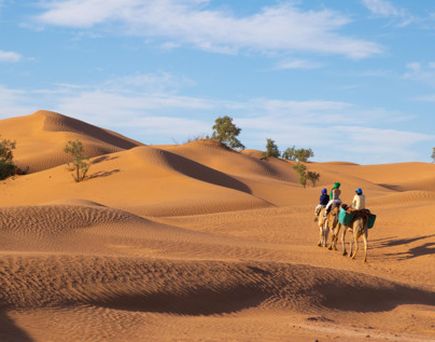 Drei Personen reiten auf Kamelen durch sandige Dünenlandschaft unter blauem Himmel in Ägypten