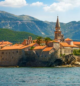 Blick auf die historische Altstadt von Budva in Montenegro mit steinernen Gebäuden, Kirchturm, Stadtmauer und Bergkulisse an der Adriaküste
