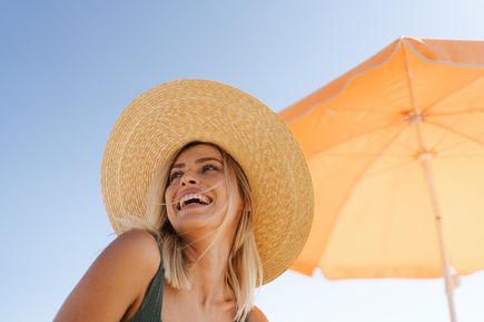 Frau mit großem, geflochtenem Strohhut sitzt unter einem orangefarbenen Sonnenschirm an einem sonnigen Tag