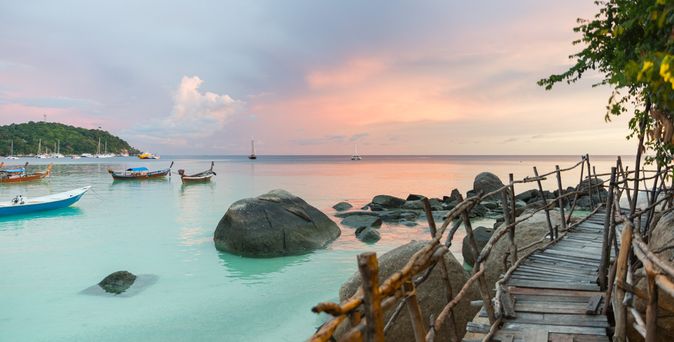 Holzsteg am Ufer mit Felsen im Wasser und mehreren Booten bei Sonnenuntergang