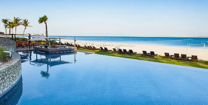 Infinity-Pool mit Blick auf den Sandstrand und das Meer, umgeben von Palmen und Liegestühlen