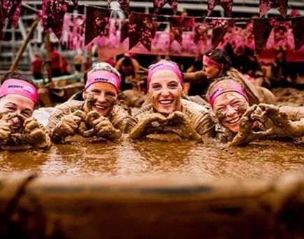 Vier Teilnehmende beim Muddy Angel Run liegen im Schlammparcours und formen mit schlammigen Händen ein Herz, im Hintergrund sind pinke Fahnen der Veranstaltung zu sehen