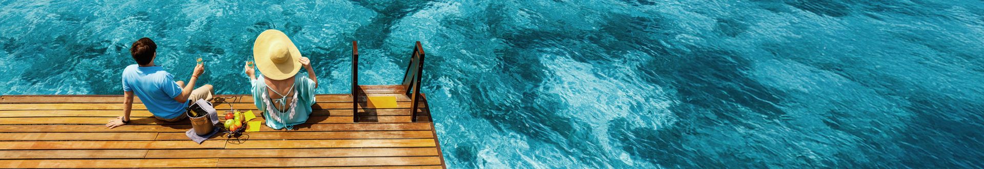 Zwei Personen sitzen entspannt am Rand eines Holzstegs über klarem türkisblauem Meer und genießen den sonnigen Ausblick