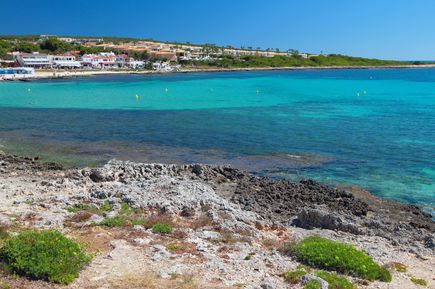 Aussicht auf die Küste von Punta Prima auf Menorca mit türkisblauem Meer, flachen Felsen im Vordergrund und Ferienhäusern sowie Hotels am Strand im Hintergrund