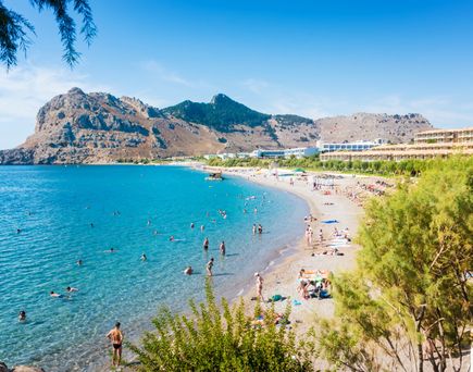 Strand mit Badegästen und Hotels vor felsigem Gebirge an der Küste von Kolymbia auf Rhodos.