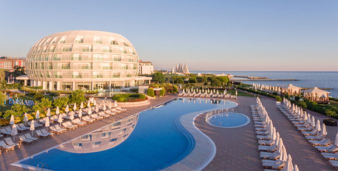 Großes, rundliches Hotelgebäude neben einem geschwungenen Außenpool mit Reihen von Liegestühlen und Sonnenschirmen am Ufer