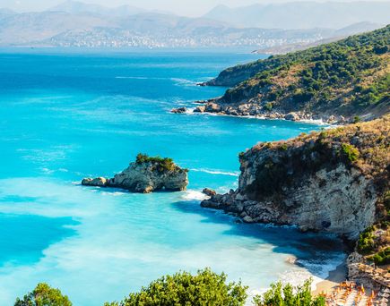 Küste mit felsigen Klippen und türkisfarbenem Meer in Albanien, im Hintergrund Berge und bewölkter Himmel