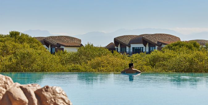 Person lehnt am Rand eines Infinity-Pools mit Blick auf grüne Büsche und runde, strohgedeckte Häuser im Hintergrund.