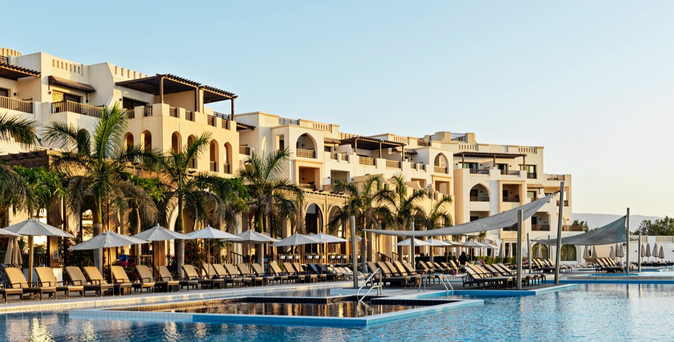 Hotel mit mehreren Stockwerken, umgeben von Palmen und einem großen Pool mit Liegen und Sonnenschirmen.