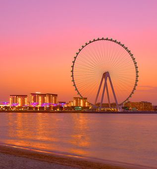 Riesenrad am Ufer mit beleuchteten Gebäuden bei Sonnenuntergang und ruhigem Wasser