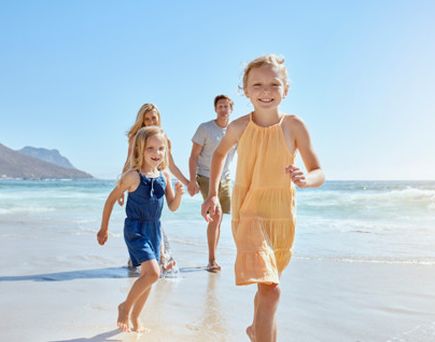 Drei Kinder laufen barfuß am Strand, zwei Mädchen vorne in blauem und gelbem Kleid, Meer und Berge im Hintergrund.