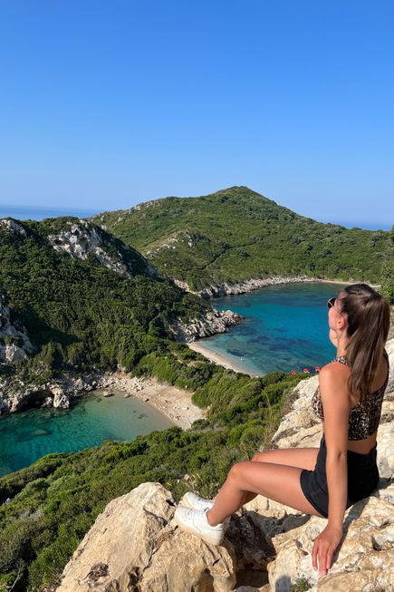 Frau in sportlicher Kleidung sitzt auf einem Felsen und blickt auf eine grüne Bucht mit klarem, blauem Wasser auf Korfu.