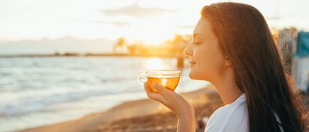 Frau genießt bei Sonnenuntergang ein warmes Getränk aus einer Glastasse am Strand
