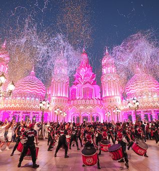 Bunte Festbeleuchtung an orientalisch gestalteten Gebäuden im Global Village Dubai, davor tanzende Menschen und Trommler, bunte Konfetti und Luftschlangen am Nachthimmel.