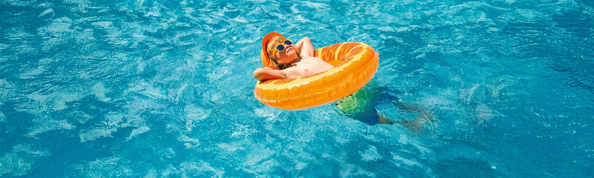 Kind mit Sonnenbrille liegt relaxed in einem Schwimmreifen im Pool, umgeben von türkisblauem Wasser