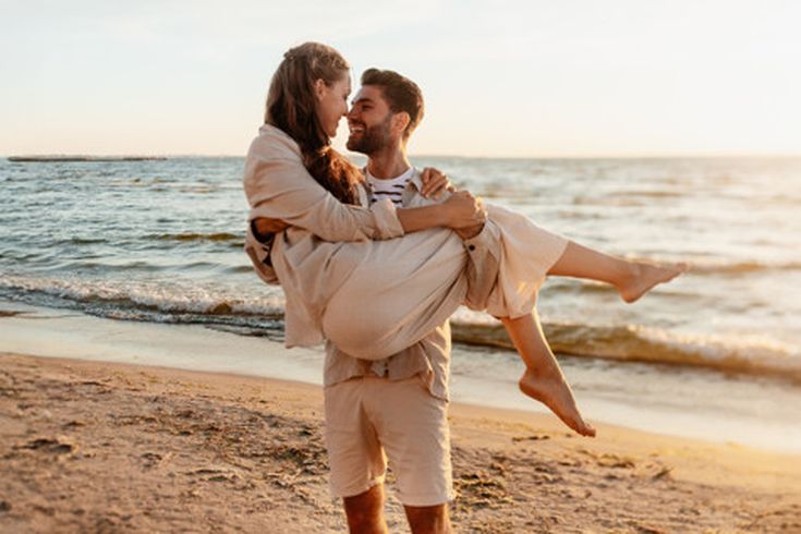 Mann trägt seine Partnerin am Strand im Sonnenuntergang, beide schauen sich verliebt an