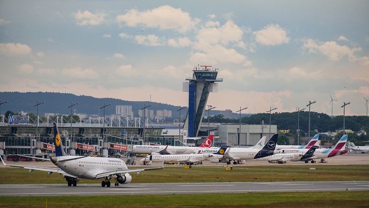 Ein Lufthansa-Flugzeug rollt auf der Start- und Landebahn des Albrecht Dürer Airport Nürnberg, während vor Tower, Terminalgebäude und mehreren Flugzeugen verschiedener Airlines reger Flugbetrieb auf dem Vorfeld zu sehen ist
