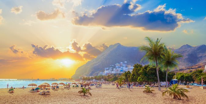 Strand mit Sonnenschirmen und Palmen, im Hintergrund ein Berg und eine Stadt, Sonnenuntergang mit Wolken und Strahlen.