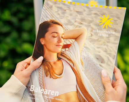 Person hält Katalog mit Titel 'Balearen' und Logo von 'Schaunisland reisen', auf dem eine Frau in weißem Top am Strand liegt.