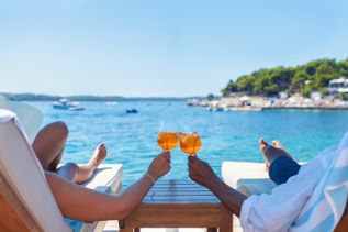 Zwei Personen stoßen mit orangenen Getränken in Gläsern an, während sie am Meer liegen, mit Blick auf Wasser und Himmel