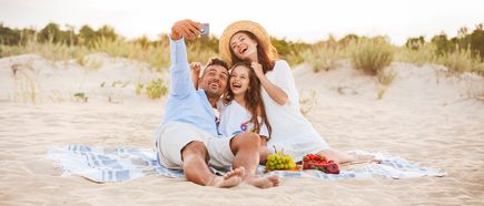 Familie sitzt auf einer Picknickdecke am Strand und macht gemeinsam ein Selfie, mit Obst, Sanddünen und sommerlicher Umgebung