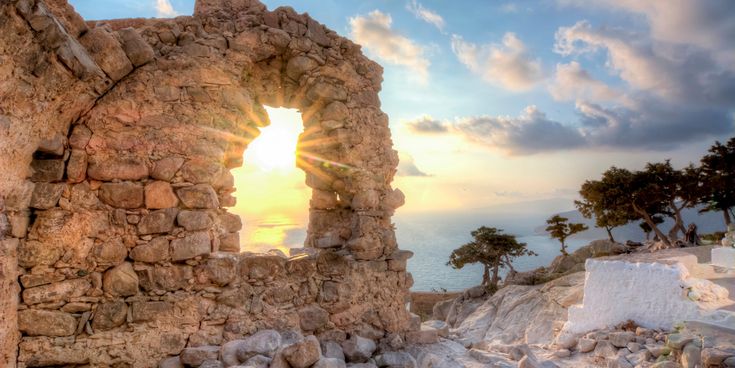 Sonnenstrahlen scheinen durch ein rundes Fenster in einer alten Steinmauer mit Blick auf das Meer und Bäume am felsigen Ufer.