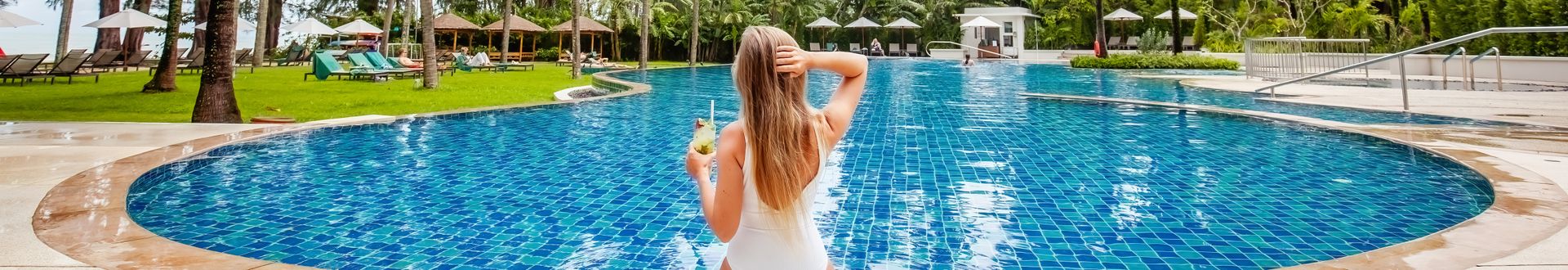 Frau in weißem Badeanzug sitzt am Rand eines großen Hotelpools und hält ein erfrischendes Getränk, umgeben von Palmen und Sonnenliegen in einer tropischen Ferienanlage