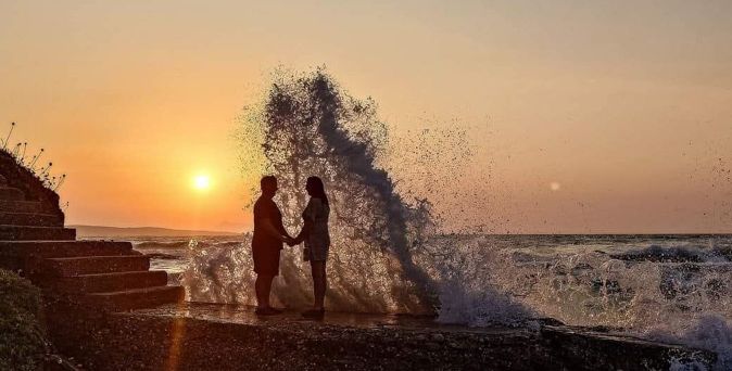 Silhouetten eines Paares, das sich am Strand bei Sonnenuntergang an den Händen hält, während eine große Welle im Hintergrund aufspritzt.