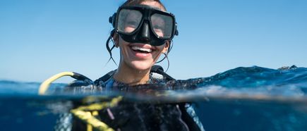 Taucherin mit schwarzem Neoprenanzug und gelbem Atemregler im offenen Meer, Wasseroberfläche halb im Bild.