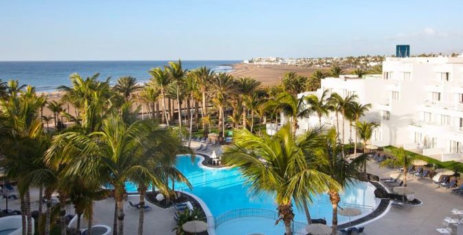 Hipotels Hotelanlage mit großem Pool, umgeben von Palmen, Liegestühlen und weißen Gebäuden, Meer im Hintergrund