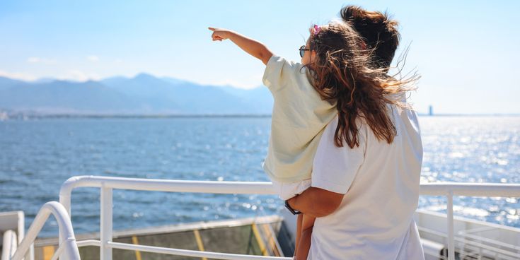 Ein Mann steht auf einem Deck mit Blick auf das Meer und hält ein Kind im Arm, das in die Ferne über das Wasser zeigt. Im Hintergrund sind Berge und ein klarer Himmel zu sehen