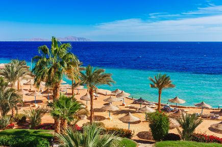 Strand mit Palmen und Sonnenschirmen am klaren Meer in Sharm el-Sheikh, Blick auf die Tiran-Insel am Horizont