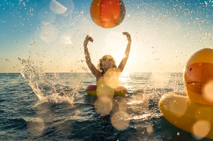 Kind spielt im Meer mit einem bunten Strandball und Schwimmring, während Wasser spritzt und die Sonne über dem Ozean steht