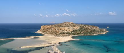 Eine sandige Landzunge verbindet den Strand mit einer flachen Felseninsel, umgeben von türkisfarbenen und dunkleren Meeresbereichen. Der Himmel ist klar und blau, und die Landschaft wirkt weit und unbebaut.