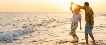 Ein Paar tanzt lachend am Meer im Sonnenuntergang, während die Wellen ihre Füße umspülen
