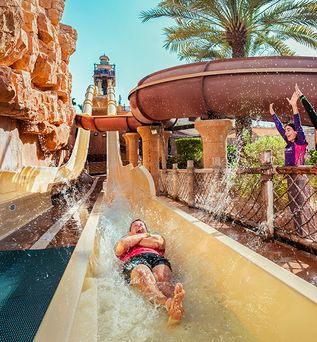 Wasserrutsche im Wild Wadi Wasserpark Dubai mit Person auf dem Rücken rutschend und zwei Personen am Rand, die die Arme heben.