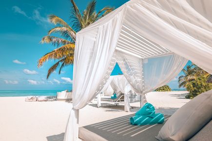 Strand mit weißen Sandliegen unter weißen Stoffdächern und Palmen im Hintergrund
