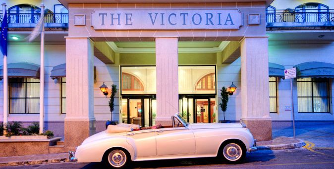 Klassischer weißer Cabrio vor dem Eingangsbereich des Hotels "The Victoria" mit Säulen und großen Fenstern.