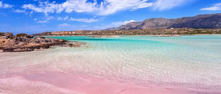 Kretas flacher Strand mit rosa schimmerndem Sand trifft auf klares, türkisfarbenes Wasser. Dahinter liegen Felsen und eine bergige Landschaft unter einem blau-weißen Himmel.