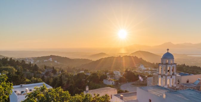 Sonnenaufgang über einer hügeligen Landschaft mit einem weißen Kirchengebäude und einer blauen Kuppel im Vordergrund.