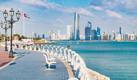 Die Corniche in Abu Dhabi mit geschwungener Uferpromenade, Sitzbänken, Flagge der Vereinigten Arabischen Emirate und moderner Skyline am Persischen Golf.