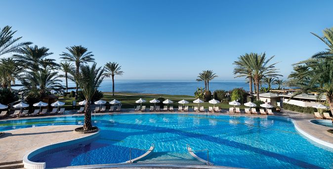 Großer, geschwungener Hotelpool mit Liegen und Sonnenschirmen, umgeben von Palmen, mit Blick auf das Meer im Hintergrund