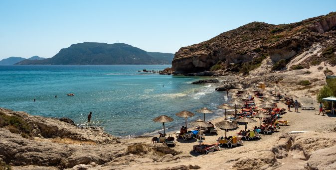 Strand mit Liegestühlen und Sonnenschirmen vor felsiger Küste und ruhigem Meer mit Bergen im Hintergrund