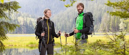 Zwei Wanderer mit Rucksäcken und Trekkingstöcken stehen lächelnd vor einer Wald- und Seenlandschaft