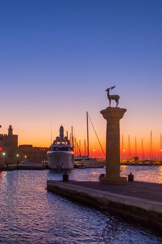 Blick auf den Mandraki-Hafen in Rhodos bei Sonnenuntergang mit zwei Säulen, von denen eine eine Hirschstatue trägt, und einem großen Schiff am Kai.