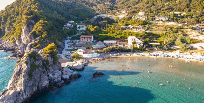 Luftaufnahme eines Strandes mit Felsen, mehreren Gebäuden und Menschen im Wasser an der Küste eines bewaldeten Hügels.